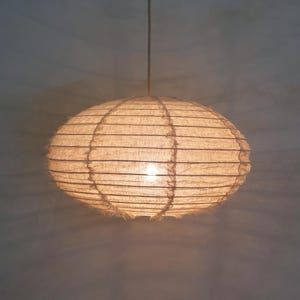 Puede incluir: Una l&aacute;mpara colgante de forma ovalada con una luz c&aacute;lida y brillante. La pantalla est&aacute; hecha de un material tejido de color natural, con l&iacute;neas horizontales y un borde deshilachado. La luz cuelga de un cable delgado.
