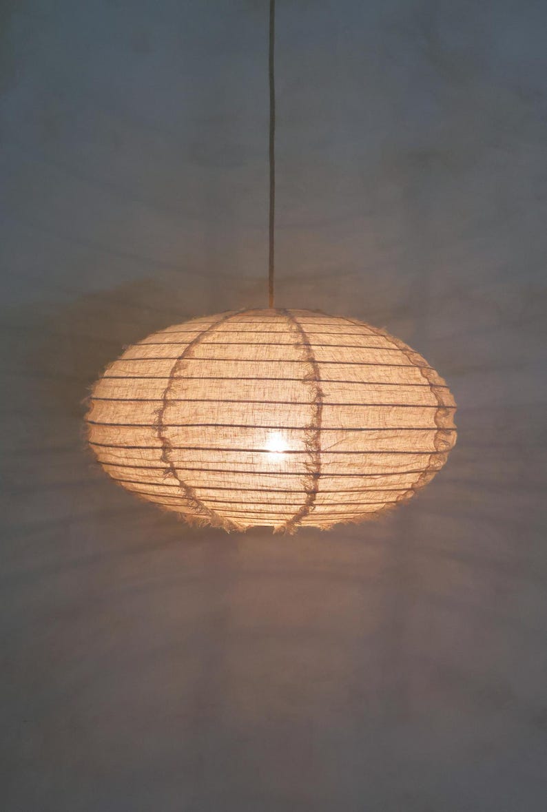 Puede incluir: Una l&aacute;mpara colgante de forma ovalada con una luz c&aacute;lida y brillante. La pantalla est&aacute; hecha de un material tejido de color natural, con l&iacute;neas horizontales y un borde deshilachado. La luz cuelga de un cable delgado.