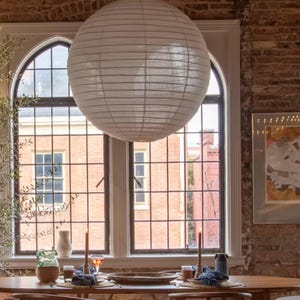 May include: A large, spherical white paper lantern pendant light with a ribbed texture, suspended from the ceiling. The light fixture features a thin, delicate paper shade that diffuses light, creating a soft and even glow throughout the room.