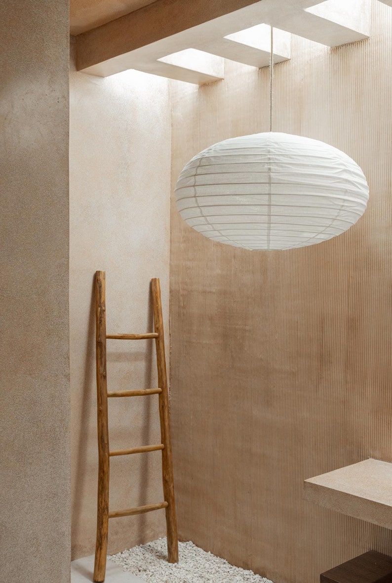 May include: A white oval paper lantern with horizontal ribbing suspended from the ceiling by a thin cord, positioned above a tall, slender wooden ladder with four rungs, featuring a natural light brown finish and a simple, rustic construction.