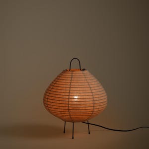 Peut inclure: Une lampe de table lanterne en papier ronde avec une lueur orange chaude, dot&eacute;e d'une fine structure en fil noir, d'une poign&eacute;e noire sur le dessus et de trois pieds fins en m&eacute;tal noir. Un cordon &eacute;lectrique noir part de la base vers la droite.