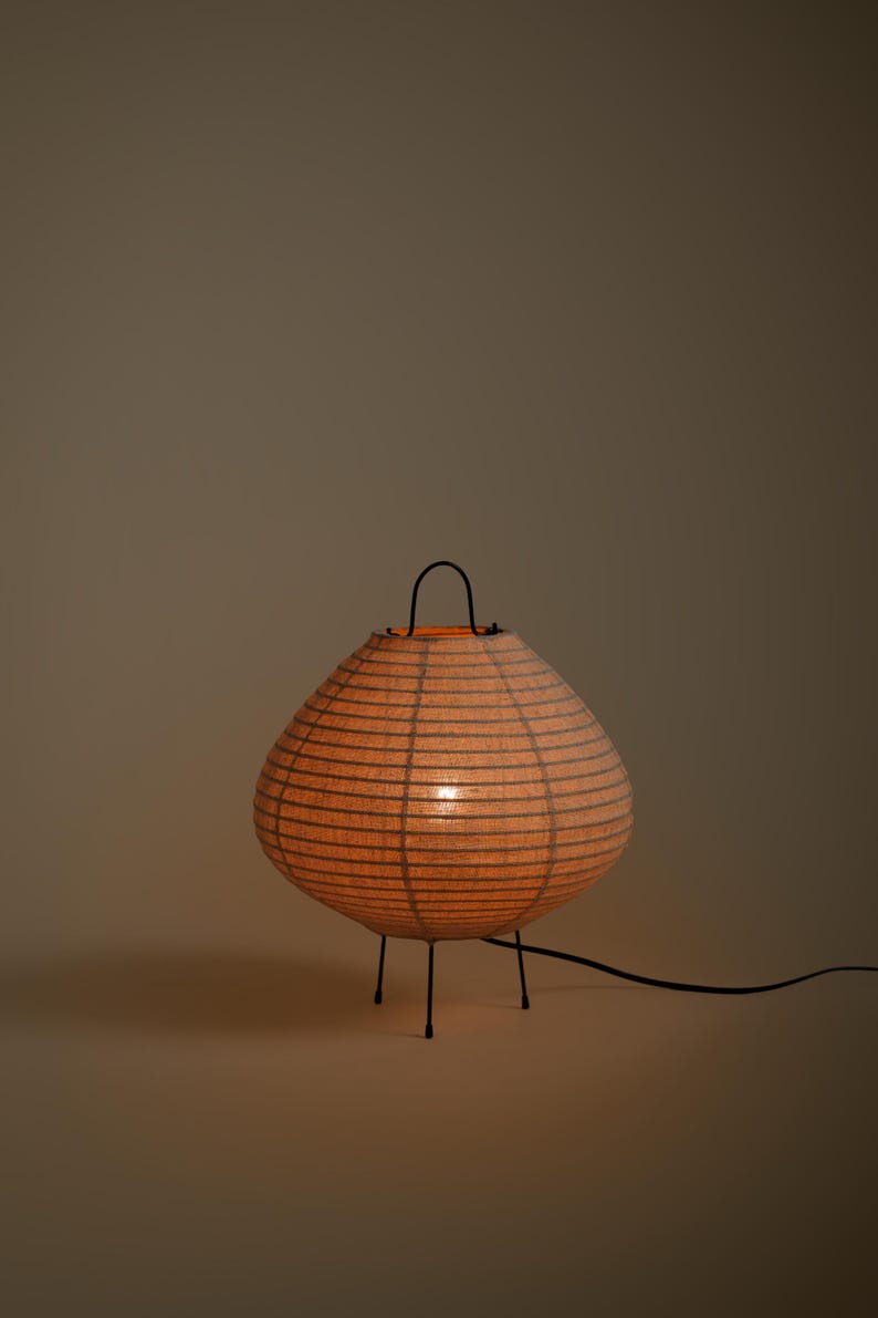 Peut inclure: Une lampe de table lanterne en papier ronde avec une lueur orange chaude, dot&eacute;e d'une fine structure en fil noir, d'une poign&eacute;e noire sur le dessus et de trois pieds fins en m&eacute;tal noir. Un cordon &eacute;lectrique noir part de la base vers la droite.