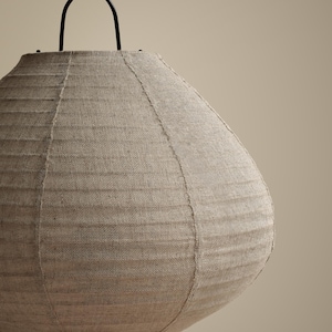 Peut inclure: Une lampe de table ronde dot&eacute;e d'un abat-jour en tissu beige textur&eacute; avec des coutures verticales visibles. La lampe est soutenue par une fine structure en m&eacute;tal noir avec une poign&eacute;e incurv&eacute;e sur le dessus et un cordon d'alimentation noir.