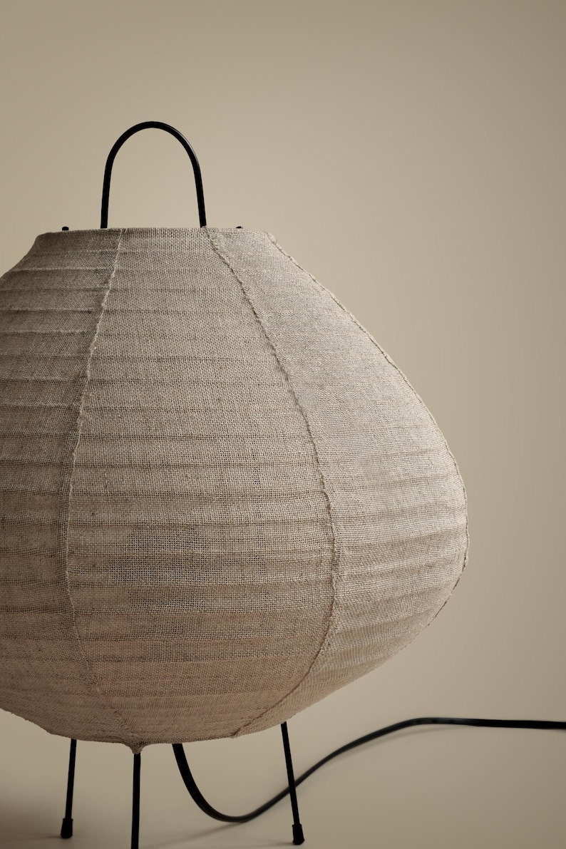 Peut inclure: Une lampe de table ronde dot&eacute;e d'un abat-jour en tissu beige textur&eacute; avec des coutures verticales visibles. La lampe est soutenue par une fine structure en m&eacute;tal noir avec une poign&eacute;e incurv&eacute;e sur le dessus et un cordon d'alimentation noir.