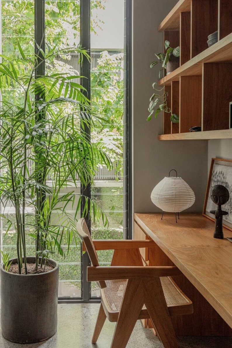 The Frank Table Lamp, a handcrafted linen lantern-style lamp with a black metal frame, sits elegantly on a wooden desk in a warm, nature-inspired home office. Its soft, diffused light complements the earthy tones and Japandi aesthetic, perfect for creating a cozy and stylish workspace.