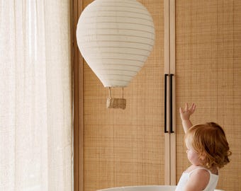 May include: A hanging hot air balloon decoration made of off-white fabric with horizontal ribbed detailing and a small woven wicker basket suspended from the bottom by thin cords, designed as a lightweight mobile for interior room decor.