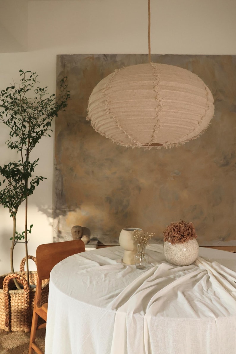 Puede incluir: Una l&aacute;mpara colgante redonda beige cuelga sobre una mesa redonda cubierta con un mantel blanco. La habitaci&oacute;n presenta una paleta de colores neutros con una gran pintura abstracta en la pared. Jarrones decorativos y una cesta de mimbre completan la decoraci&oacute;n.