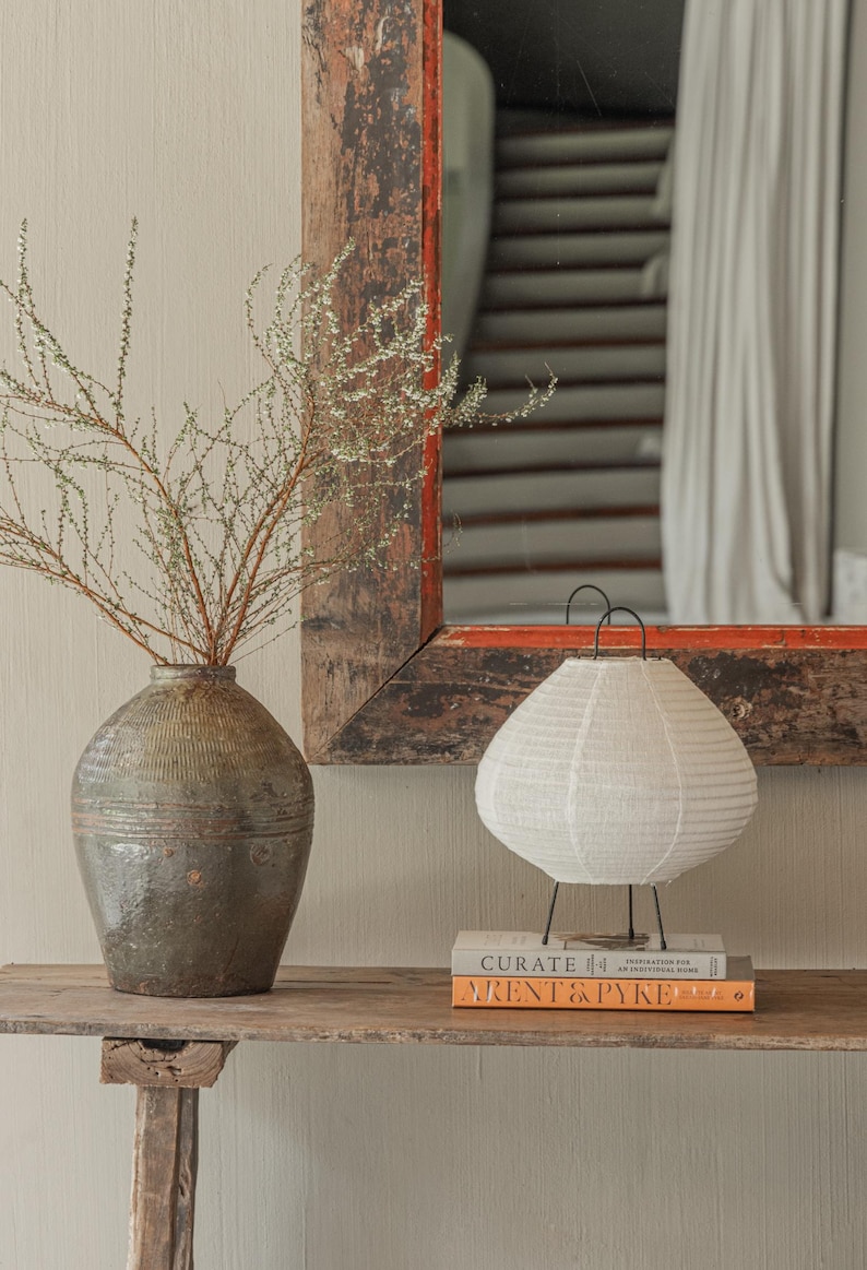 A beautifully styled boho-chic interior featuring a handcrafted linen table lamp with a soft, organic glow. The lamp sits atop two stacked design books on a rustic wooden console table, accompanied by an artisanal textured vase with delicate branches. A vintage wooden mirror with distressed edges reflects a cozy, neutral-toned space in the background. The scene exudes warmth, simplicity, and timeless craftsmanship.