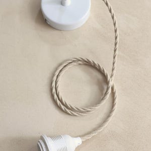 May include: A white ceiling canopy with a matching light socket and a beige twisted fabric cord. The cord is looped and connected to the canopy and socket, creating a simple, modern design for a pendant light.