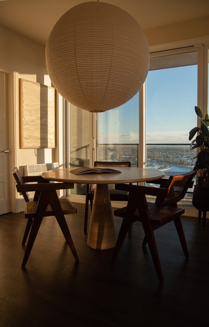 May include: A modern dining room set with a round wooden table and four chairs. A large paper lantern hangs above the table. The room has large windows with a view of the city.