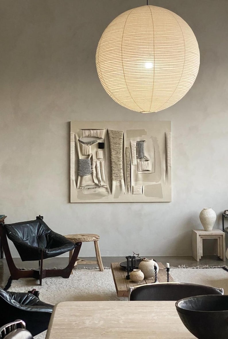 A serene, modern living space featuring the Empire Shade – a handcrafted, off-white linen pendant light with delicate horizontal lines. The Japandi-inspired room blends neutral tones, textured wall art, and organic materials, with a cozy mix of black leather lounge chairs, a rustic wooden coffee table, and ceramic decor. A perfect balance of warmth, texture, and contemporary elegance.
