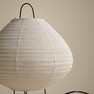 Peut inclure: Une lampe de table ronde dot&eacute;e d'un abat-jour en tissu blanc cass&eacute; textur&eacute; avec des d&eacute;tails nervur&eacute;s horizontaux. La lampe est soutenue par de fines pattes en m&eacute;tal noir et comprend une poign&eacute;e en m&eacute;tal noir sur le dessus, avec un cordon &eacute;lectrique noir.