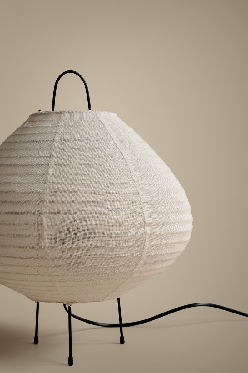 Peut inclure: Une lampe de table ronde dot&eacute;e d'un abat-jour en tissu blanc cass&eacute; textur&eacute; avec des d&eacute;tails nervur&eacute;s horizontaux. La lampe est soutenue par de fines pattes en m&eacute;tal noir et comprend une poign&eacute;e en m&eacute;tal noir sur le dessus, avec un cordon &eacute;lectrique noir.