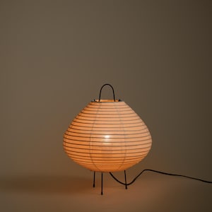 Peut inclure: Une lampe lanterne en papier ronde avec des d&eacute;tails nervur&eacute;s horizontaux, &eacute;mettant une lueur chaude. La lampe pr&eacute;sente une structure en m&eacute;tal noir avec trois pieds fins et une poign&eacute;e incurv&eacute;e sur le dessus, reli&eacute;e &agrave; un cordon &eacute;lectrique noir.