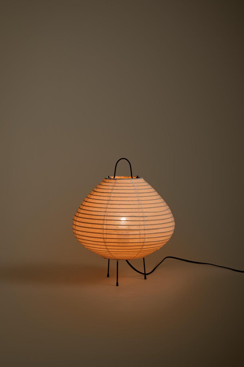 Peut inclure: Une lampe lanterne en papier ronde avec des d&eacute;tails nervur&eacute;s horizontaux, &eacute;mettant une lueur chaude. La lampe pr&eacute;sente une structure en m&eacute;tal noir avec trois pieds fins et une poign&eacute;e incurv&eacute;e sur le dessus, reli&eacute;e &agrave; un cordon &eacute;lectrique noir.