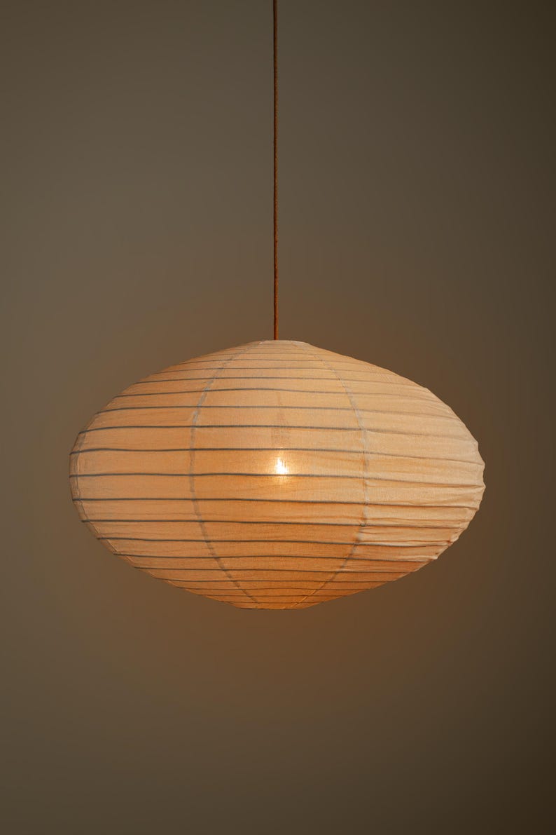 May include: A round, off-white paper pendant lamp with a ribbed texture and horizontal wire structure. The light fixture is suspended by a thin, dark brown cord, emitting a soft, warm glow from the center, creating a diffused lighting effect.