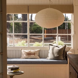Peut inclure: Une suspension en papier blanc est suspendue au-dessus d'une banquette intégrée dotée d'un coussin rayé bleu et blanc, d'un coussin rectangulaire avec une illustration de homard brun et d'un coussin carré gris uni à bords effilochés près d'une table.