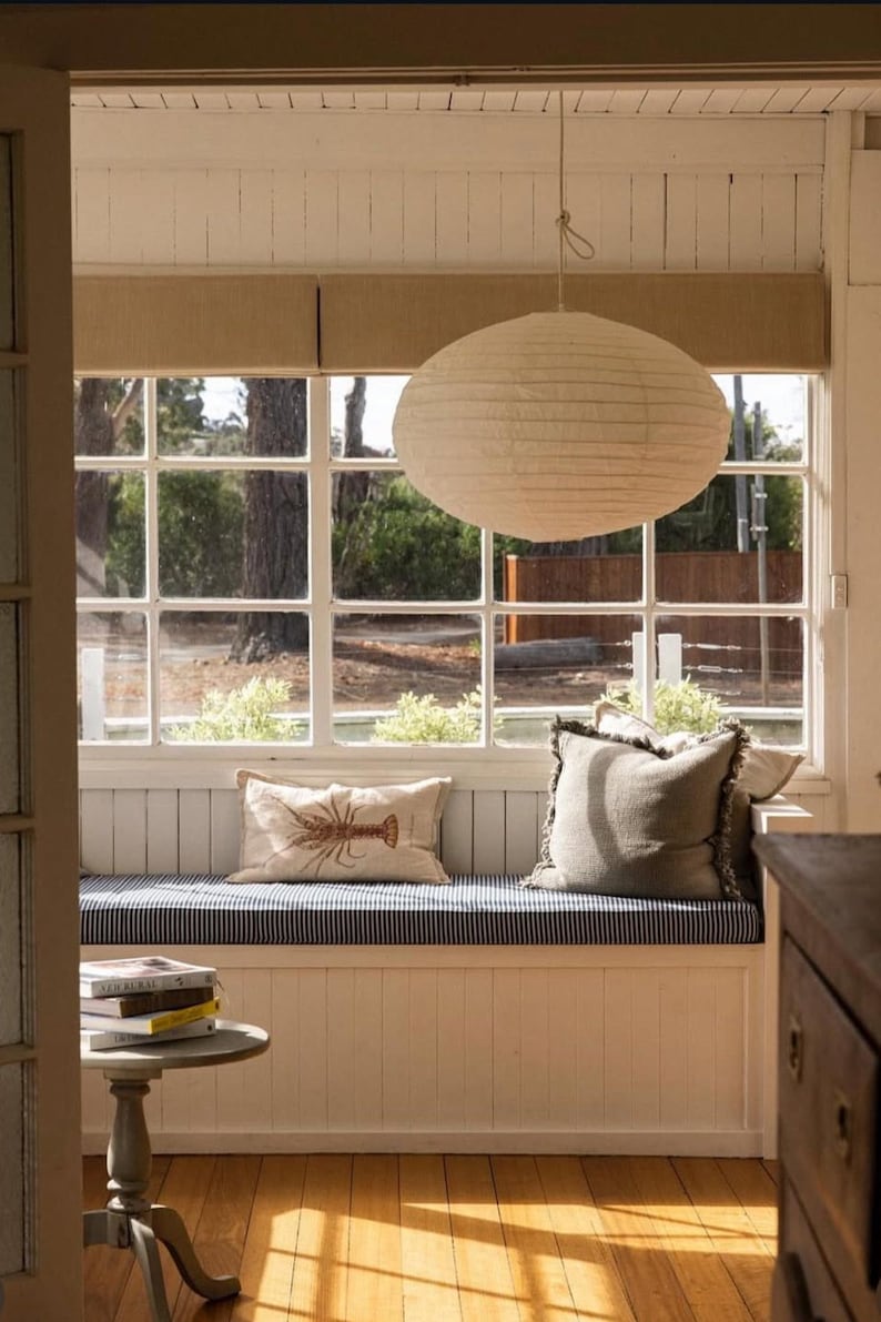 May include: A white paper pendant light hangs above a window seat with a blue and white striped cushion. The seat features a rectangular beige pillow with a brown lobster print and a square gray fringed pillow. A small round table holds books.