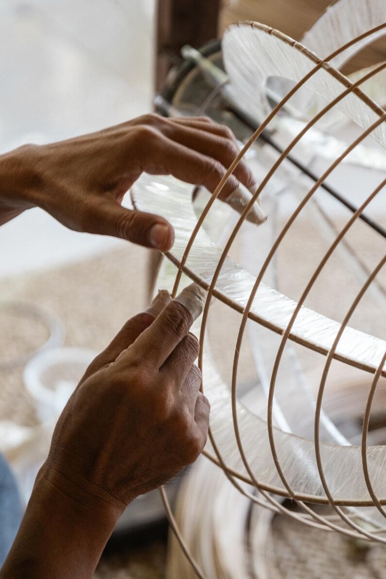 Peut inclure: Une structure sph&eacute;rique compos&eacute;e de fines nervures en bois clair dispos&eacute;es en grille, avec un mat&eacute;riau fibreux translucide et p&acirc;le superpos&eacute; entre les lattes de bois incurv&eacute;es pour former un objet arrondi en forme de cage.