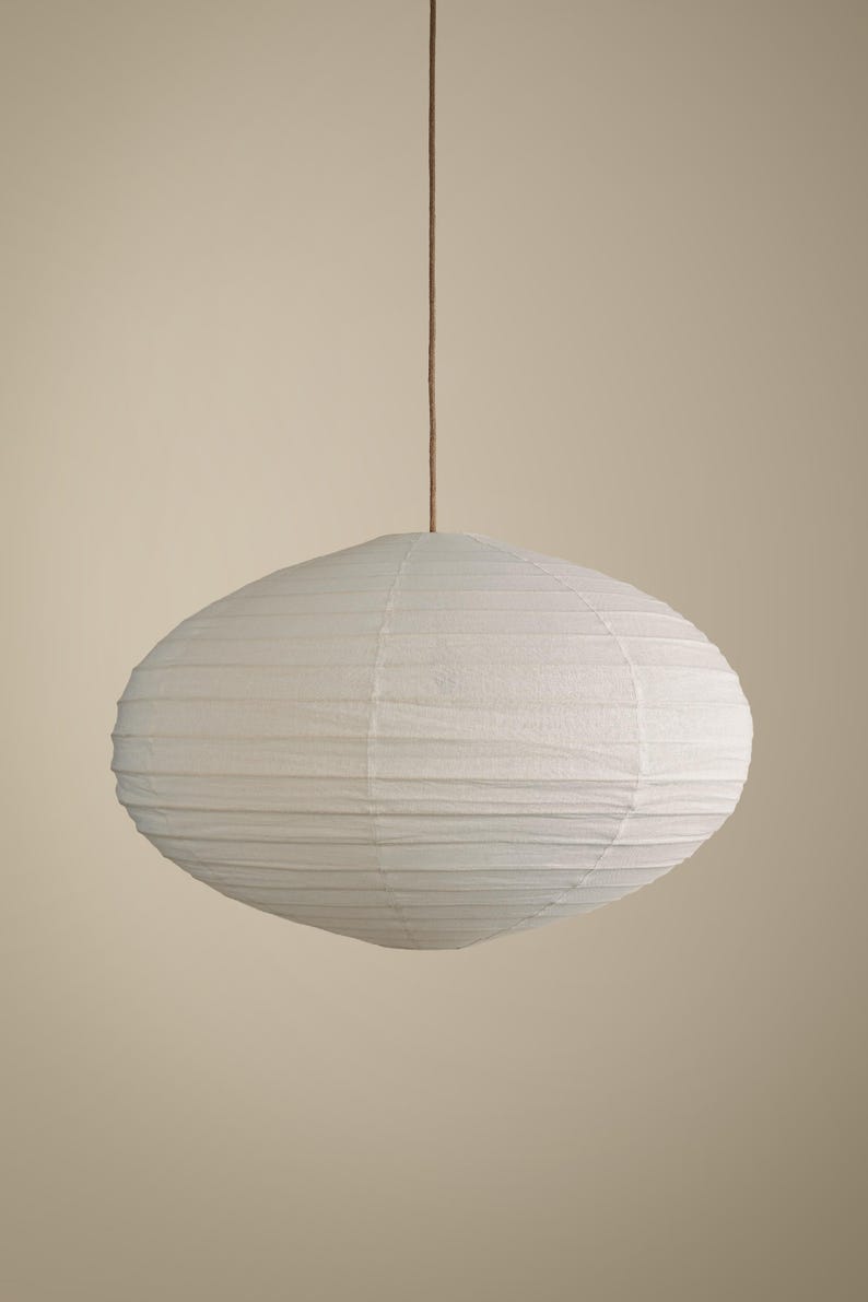 May include: A white oval-shaped paper lantern pendant light with a ribbed texture, suspended from a thin brown cord. The light fixture features a delicate, lightweight paper shade that creates a soft, rounded silhouette against a neutral background.