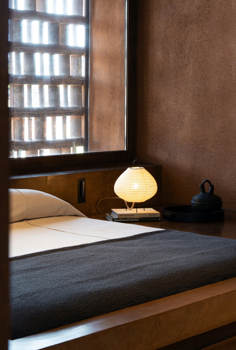 Peut inclure: Une chambre avec un lit recouvert d'une couverture textur&eacute;e gris fonc&eacute; et de draps blancs. Une lampe lanterne en papier blanc cass&eacute; repose sur une pile de livres &agrave; c&ocirc;t&eacute; d'un plateau circulaire noir avec un r&eacute;cipient &agrave; couvercle, pr&egrave;s d'une fen&ecirc;tre.