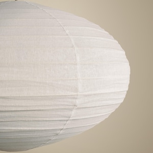 May include: A round, off-white paper lantern with a textured, ribbed surface and a visible vertical seam. The lantern is suspended from a thin, brown cord attached to the top center, creating a soft, rounded shape with horizontal ridges.