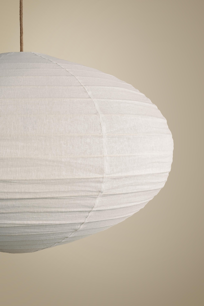 May include: A round, off-white paper lantern with a textured, ribbed surface and a visible vertical seam. The lantern is suspended from a thin, brown cord attached to the top center, creating a soft, rounded shape with horizontal ridges.