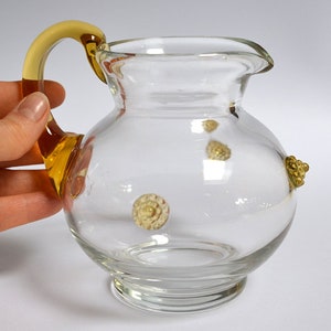 Könnte beinhalten: Ein klarer Glaskrug mit rundem Körper und ausgestelltem Rand. Er hat einen gelb-braunen Griff und dekorative goldfarbene Verzierungen. Der Krug ist etwa 13 cm hoch.