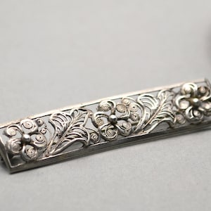 Könnte beinhalten: Eine Brosche aus Silberfiligran mit floralem Design. Die Brosche ist rechteckig und zeigt drei Blumen mit filigranen Details.