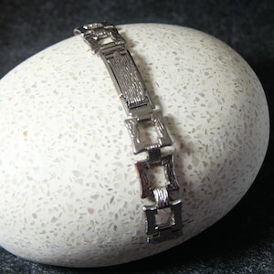 Könnte beinhalten: Ein silberfarbenes Armband mit einem strukturierten rechteckigen Mittelglied und quadratischen Gliedern. Das Armband wird auf einer weißen, gesprenkelten Oberfläche mit dunklem Hintergrund präsentiert.