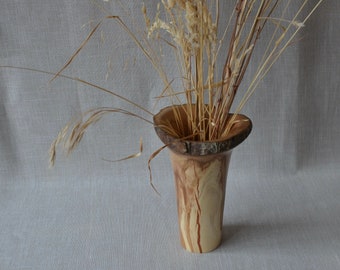 May include: A wooden vase with a natural bark rim and a light brown, cream, and reddish-brown wood grain pattern. The vase is filled with dried grasses.