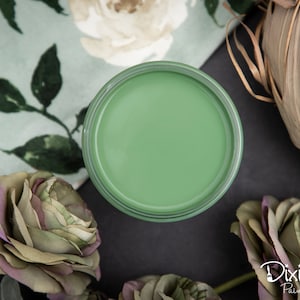 May include: A small jar of green paint with a white label that reads "Dixie Belle Paint Company". The jar is sitting on a black surface with a floral fabric and a woven basket in the background.