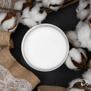 Puede incluir: Una pintura blanca en un recipiente redondo con una tapa blanca. El recipiente está rodeado de bolas de algodón y un trozo de tela de arpillera con encaje blanco.