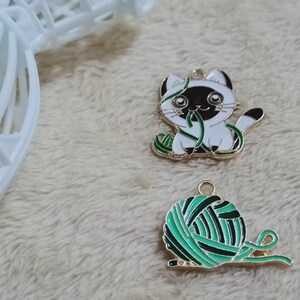 May include: Two gold-colored metal charms, one depicting a white cat with black ears and a green ball of yarn, the other a green ball of yarn with a gold-colored string.