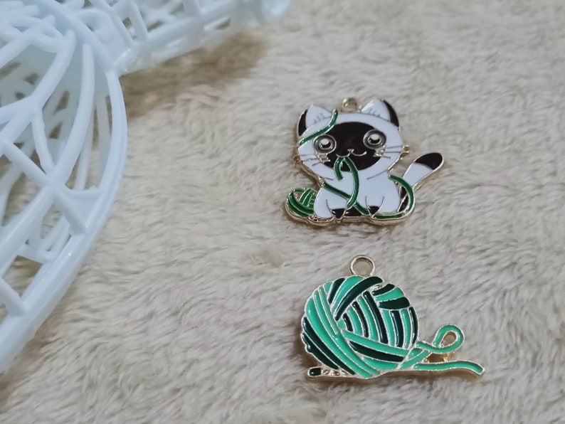 May include: Two gold-colored metal charms, one depicting a white cat with black ears and a green ball of yarn, the other a green ball of yarn with a gold-colored string.