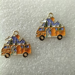May include: Two gold-colored metal charms shaped like a vintage camper van with a mountain scene on top. The camper vans are orange with white trim and blue mountains. The text "Let's Go" is on the side of the van.