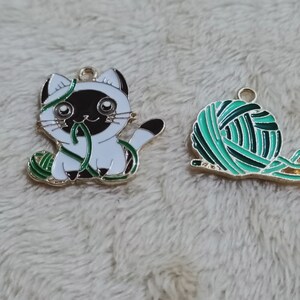 May include: Two gold metal charms, one featuring a white cat with black ears and a green ball of yarn, the other a green ball of yarn with a gold string.
