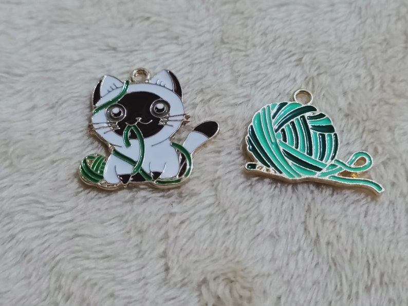 May include: Two gold metal charms, one featuring a white cat with black ears and a green ball of yarn, the other a green ball of yarn with a gold string.