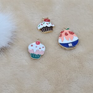 May include: Three gold-tone metal charms featuring a pink and green cupcake, a brown cupcake with red and green sprinkles, and a blue and pink shaved ice dessert with strawberries.