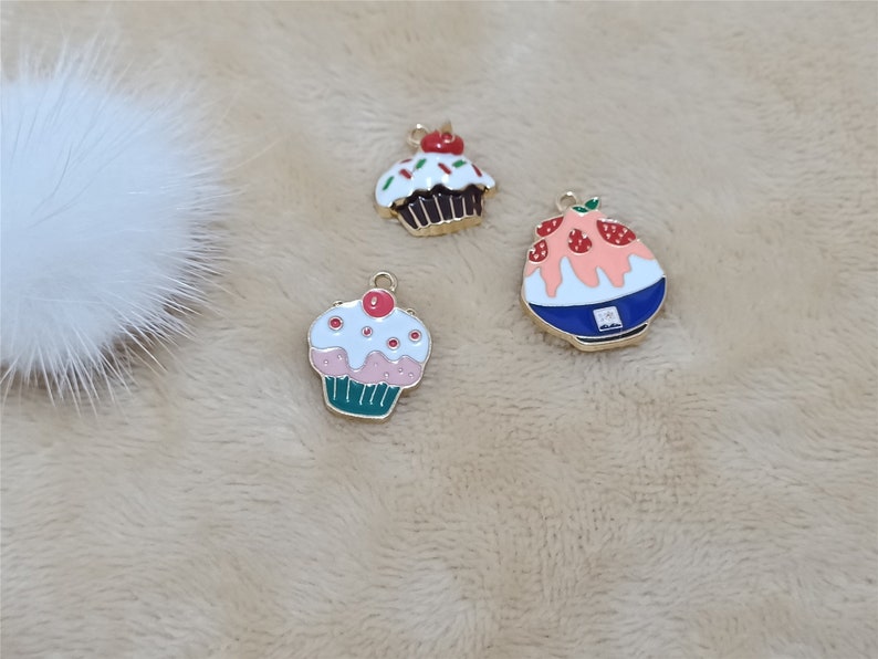 May include: Three gold-tone metal charms featuring a pink and green cupcake, a brown cupcake with red and green sprinkles, and a blue and pink shaved ice dessert with strawberries.