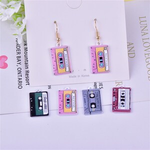 May include: A set of six colorful cassette tape earrings. The earrings are made of metal and have a gold-colored hook. The cassette tapes are pink, yellow, green, gray, and red. The cassette tapes have the text "Best of the 40's" on them.