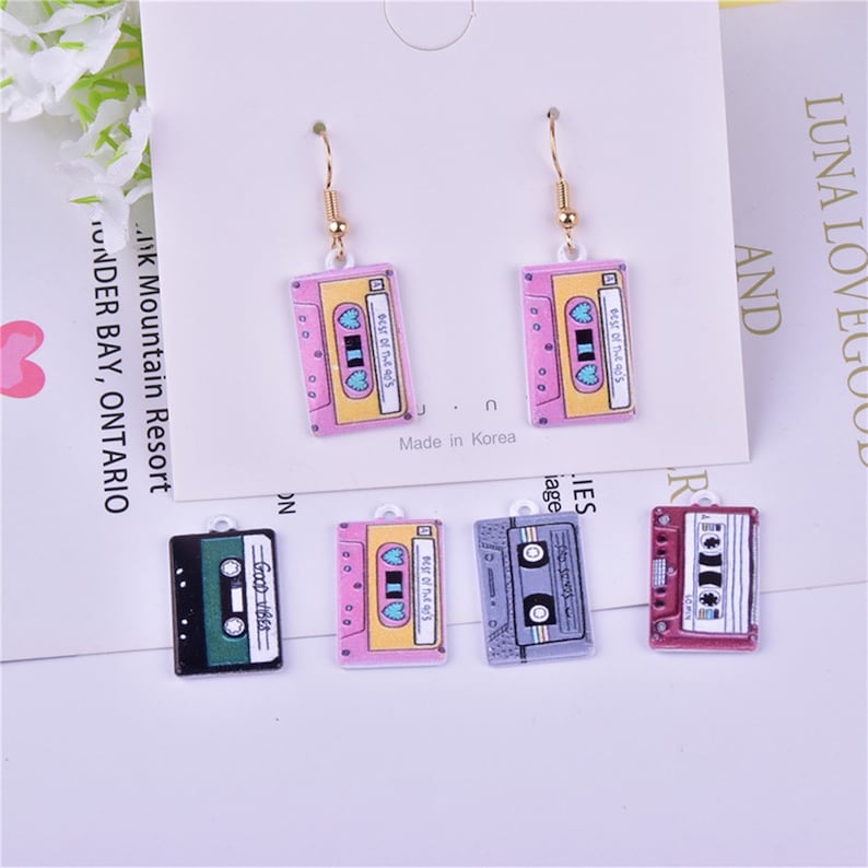 May include: A set of six colorful cassette tape earrings. The earrings are made of metal and have a gold-colored hook. The cassette tapes are pink, yellow, green, gray, and red. The cassette tapes have the text "Best of the 40's" on them.
