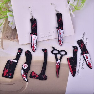 May include: A set of six black and red earrings shaped like knives, scissors, and an axe. The earrings are made of metal and have a bloody design. The earrings are made in Korea.