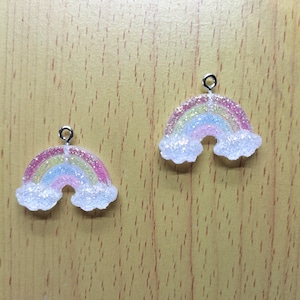 Puede incluir: Dos encantos de resina de color arcoíris con purpurina y un lazo plateado para colgar. Los encantos tienen forma de arcoíris con nubes a cada lado.
