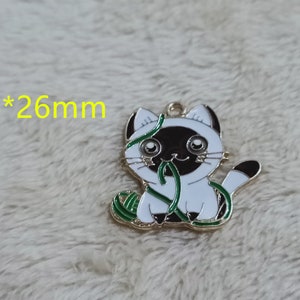 May include: Gold-tone metal charm featuring a white cat with black ears and a black face, holding a green yarn ball. The charm measures 26 mm by 26 mm.