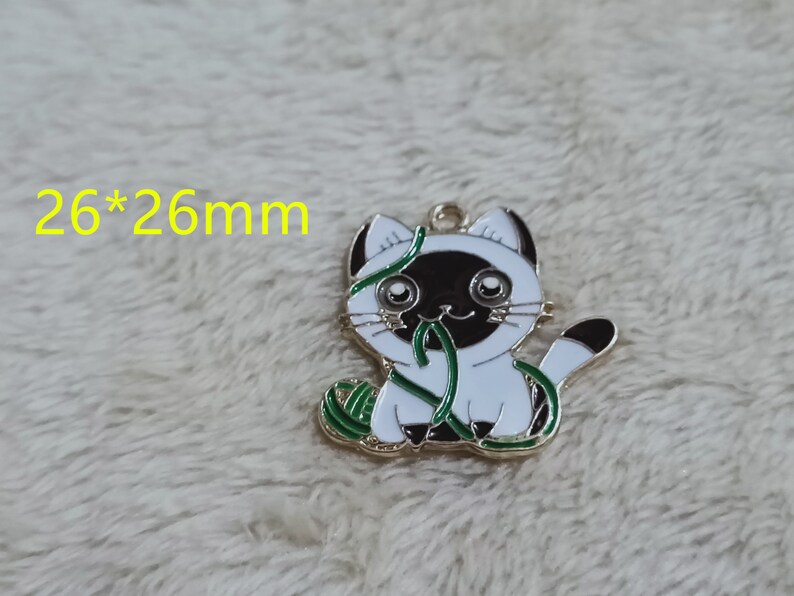 May include: Gold-tone metal charm featuring a white cat with black ears and a black face, holding a green yarn ball. The charm measures 26 mm by 26 mm.