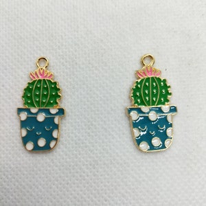 Puede incluir: Dos abalorios de metal dorado en forma de cactus en maceta con cactus verdes, flores rosas y macetas con lunares blancos.
