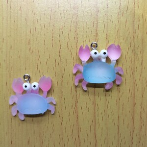 May include: Two blue and pink plastic crab charms with white eyes. The crabs have a silver loop for hanging.