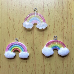Puede incluir: Tres colgantes en forma de arcoíris con detalles de purpurina y lazos plateados para colgar. Los arcoíris presentan un espectro de colores, incluyendo rosa, amarillo, azul y verde, con nubes blancas brillantes en la base. Los colgantes están sobre un fondo de madera.