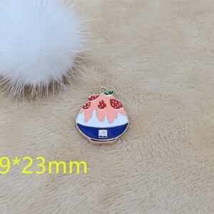 May include: A gold-toned metal charm shaped like a bowl of shaved ice with pink and white frosting and red strawberries. The charm is 19 mm by 23 mm.